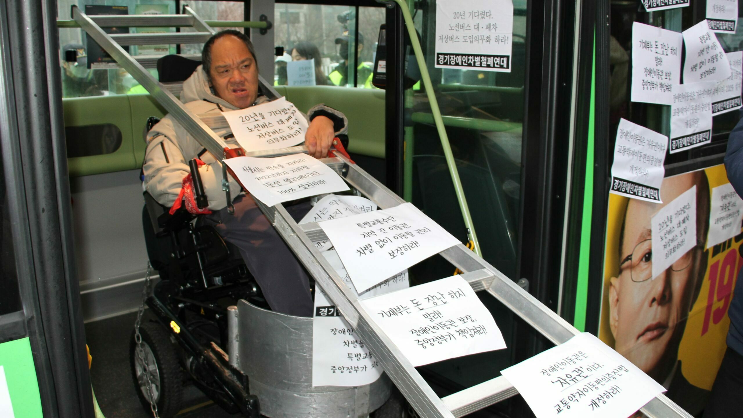 휠체어를 탄 장애인이 쇠사슬을 묶은채로 버스에 타있다. 목에는 사다리가 걸려있다. 사다리에는 20년을 기다렸다. 노선버스 대폐차시 저상버스 도입 의무화하라는 피켓이 걸려있다